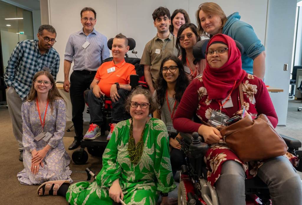 A photo of 11 of us at our Unconference in July 2024. Two of us are sat on the floor, and two people are sat in our wheelchairs. Then people are stood behind and around us seated. 